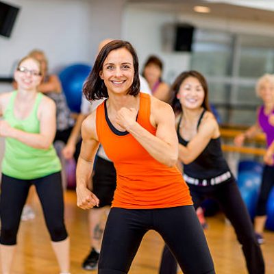 A woman is leading a multi-ethnic aerobic fitness class in an indoor gym studio - they are smiling and looking at the camera.